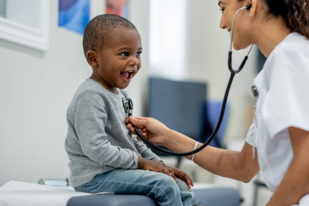 A young boy of African decent, sits up on an exam table as a female Paediatrician preforms a check-up on him. The boy is dressed casually and smiling as the doctor listens to his heart.